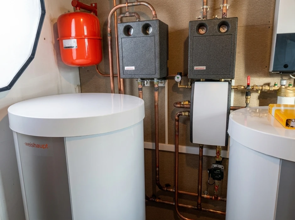 Chaufferie résidentielle moderne avec deux ballons tampons blancs **Weishaupt**, un vase d'expansion rouge, deux modules hydrauliques en mousse grise, réseau de tuyauterie en cuivre, circulateur, vannes laiton et manomètres