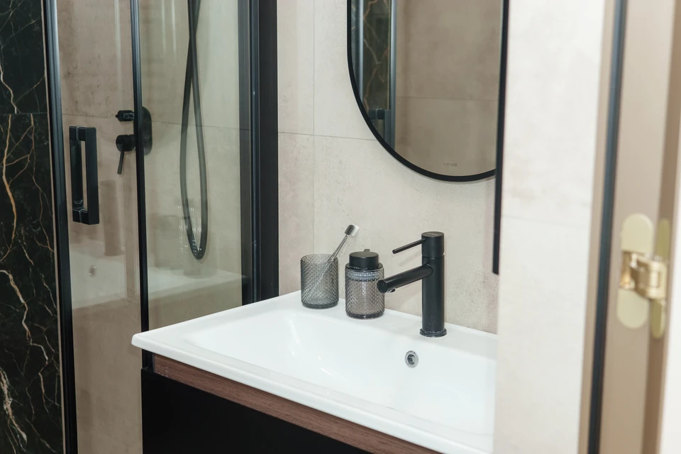 Un plombier en combinaison bleue et gants de protection installe ou répare un robinet de baignoire chromé dans une salle de bain moderne aux murs carrelés blancs
