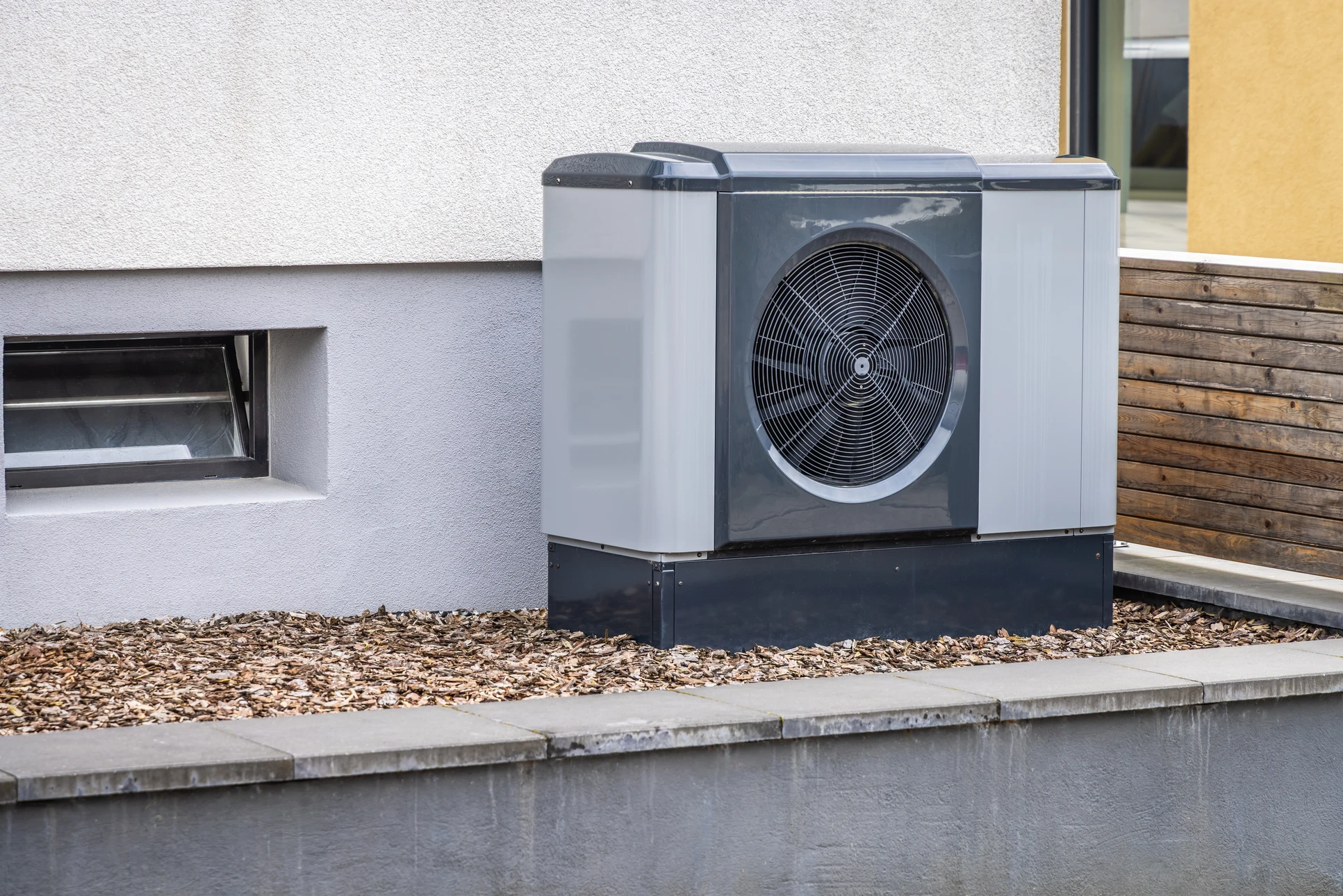 Pompe à chaleur air/eau monobloc installée en extérieur sur palette bois, devant une ferme en briques rouges et pierres calcaires